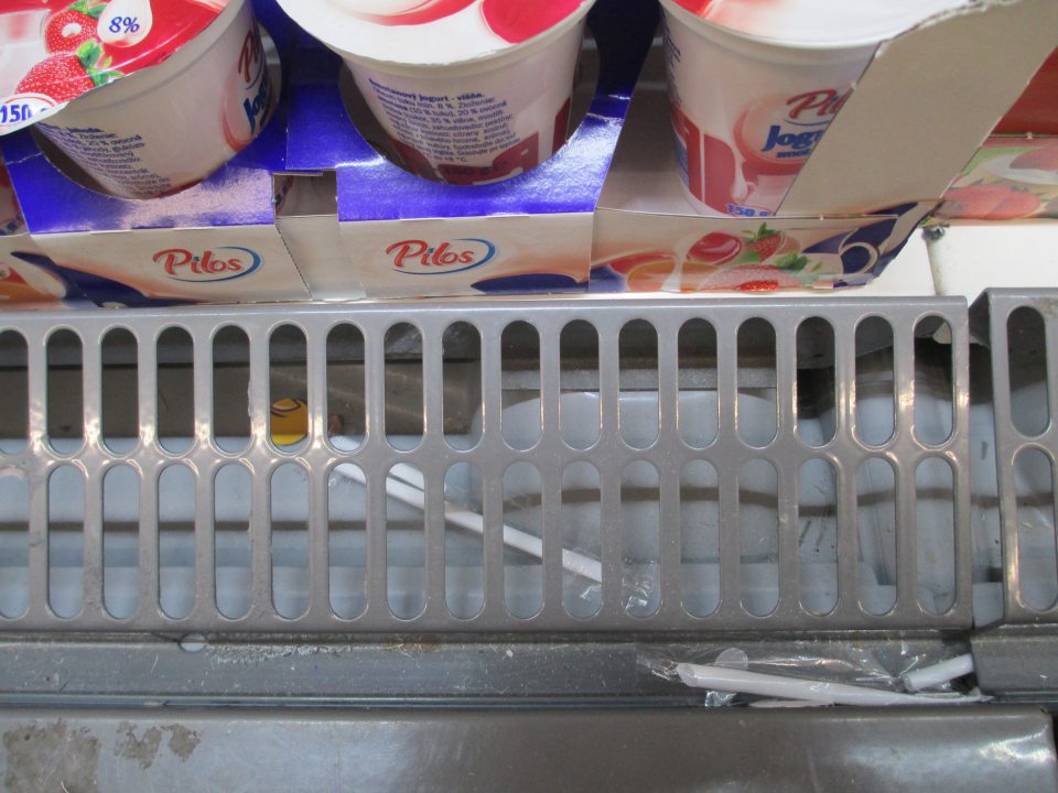 Znečistená kovová mriežka a priestor pod mriežkou na chladiacej vitríne s jogurtami.