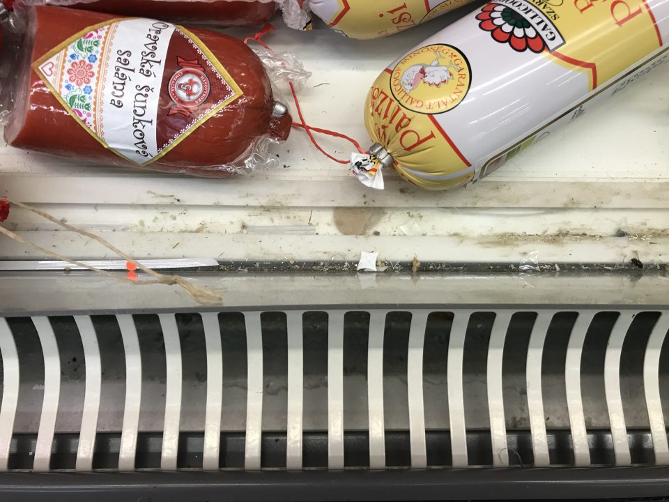 Znečistené a čiastočne poškodené plastové lišty a priestor pod nimi pri mriežkach na chladiacej vitríne s mäsovými výrobkami na predajni 