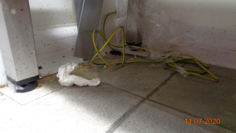 V chladiacom boxe mliečnych výrobkov podlaha pod regálmi znečistená zvyškami obalov a prachom, okrajové lišty medzi podlahou a stenou znečistené čiernou špinou.