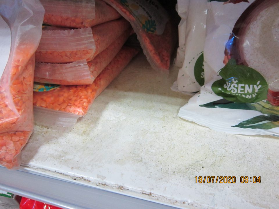 Regál na predajnej ploche, na ktorom sú umiestnené balené suché strukoviny, strúhaný kokos, znečistený nánosmi zaschnutej špiny.