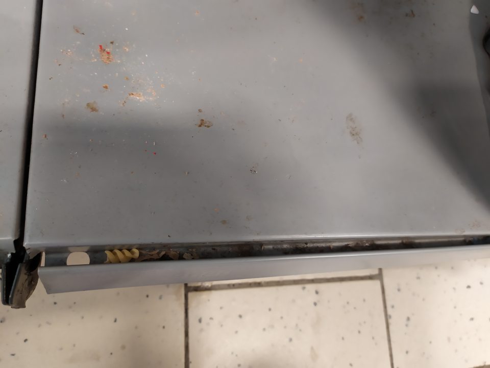 Znečistený regál a a drážka za cenovkami drobnými nečistotami a zvyškami potravín na predajnej ploche. 