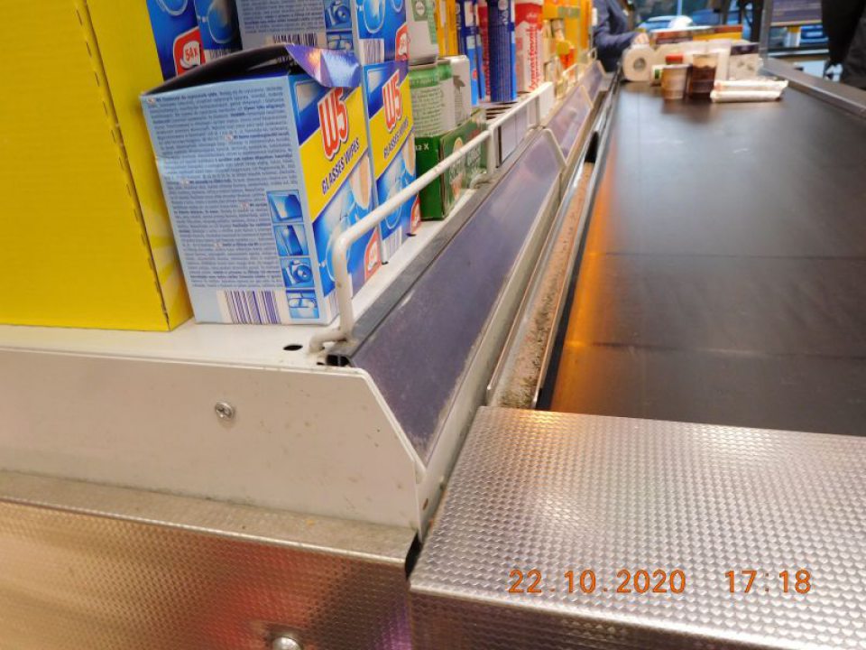 V priestore pokladní kovové koľajnice popri pohyblivom páse na ukladanie potravín znečistené hrubými nečistotami a prachom.