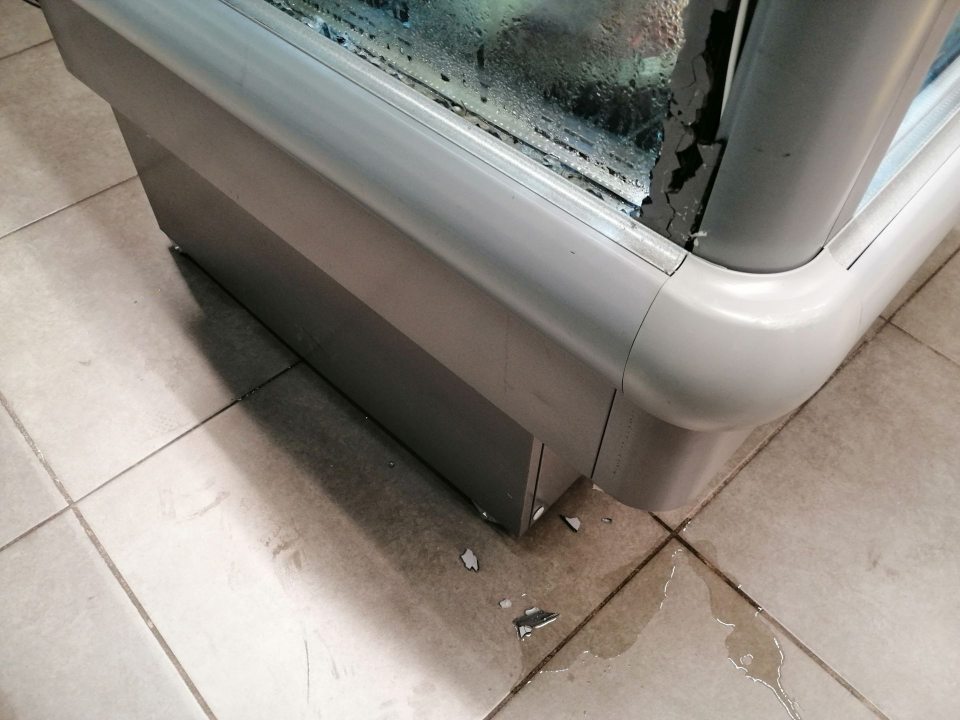 Rozbitá vonkajšia bočná stena s úlomkami skla na podlahe a stekajúcim kondenzátom mraziacej vane s ponukou nanukov pri pokladniach.