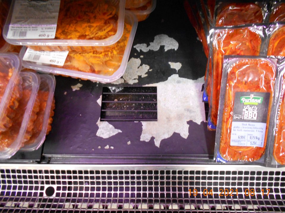 Zistený olupujúci sa náter kovového dna chladiarenskej vitríny s uchovávanými mäsovými výrobkami na predajnej ploche.
