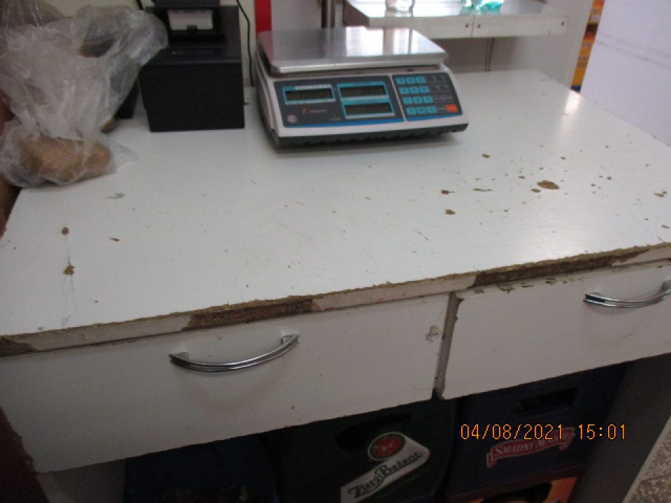 Pracovný stôl používaný na váženie oz a na prácu so zeleninou a ovocím, má na pracovnej doske poškodený povrch( drobné výmole, poškodenia zasahujúce do drevotriesky) na viacerých miestach, na prednej a bočnej hrane uvedenej pracovnej dosky je taktiež poškodený, opotrebovaný povrch lišty.