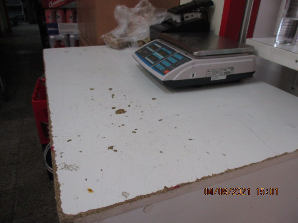 Pracovný stôl používaný na váženie oz a na prácu so zeleninou a ovocím, má na pracovnej doske poškodený povrch( drobné výmole, poškodenia zasahujúce do drevotriesky) na viacerých miestach, na prednej a bočnej hrane uvedenej pracovnej dosky je taktiež poškodený, opotrebovaný povrch lišty