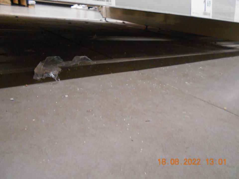 istená znečistená podlaha na predajnej ploche pod regálmi na viacerých miestach, a to zhlukmi prachu, papierikmi, odrobinkami staršieho dáta a inými nečistotami.