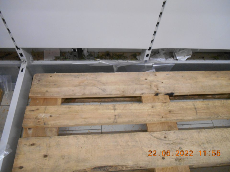 Znečistený priestor za regálom na uloženie zeleniny listami a vňaťou z uloženej zeleniny, zvyškami potrhaných fólií a kúskami papiera. 