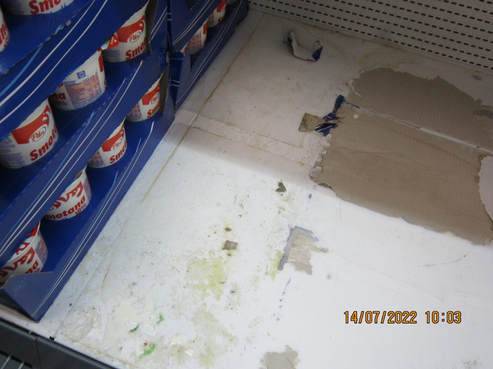 Znečistený regál v chladiacej vitríne na uchovávanie čerstvého mlieka a mliečnych výrobkov – rozliate mlieko, zaschnuté nečistoty, prilepené kúsky kartónového papiera.