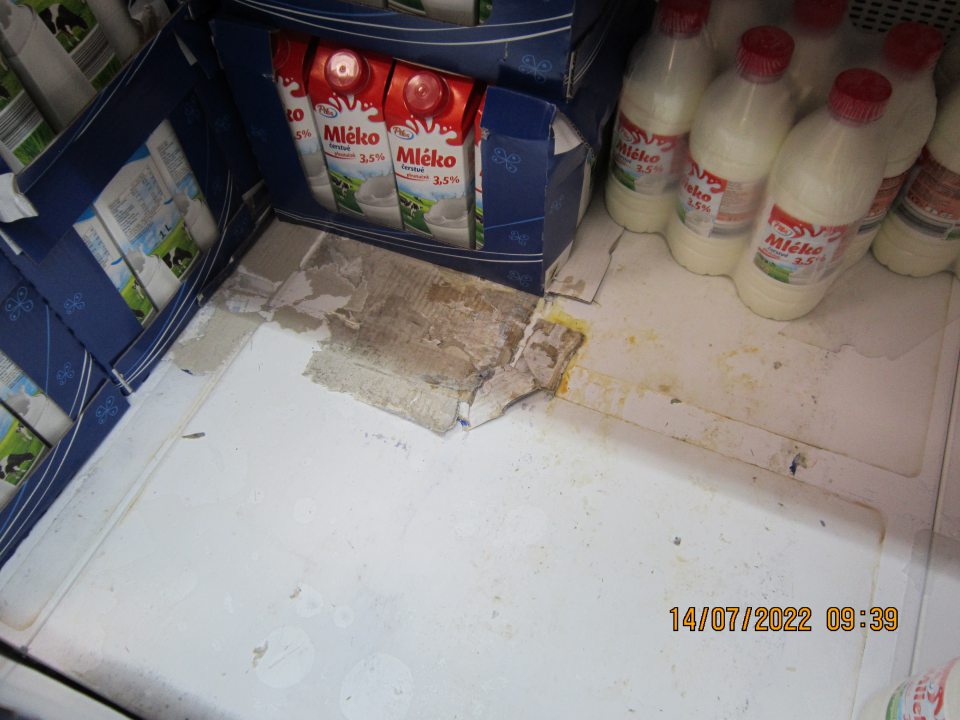 Znečistený regál v chladiacej vitríne na uchovávanie čerstvého mlieka a mliečnych výrobkov – rozliate mlieko, zaschnuté nečistoty, prilepené kúsky kartónového papiera.