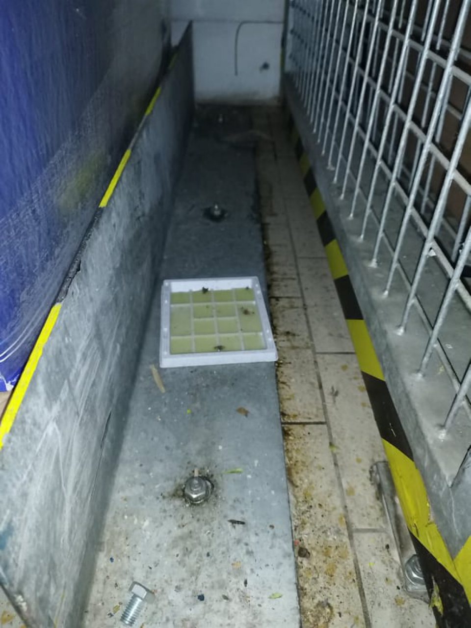 znečistená podlaha skladu medzi regálmi, drevenými paletami, kovovými spojmi a kovovými zábranami a to chumáčmi prachu, zašlou špinou, obalovým materiálom a zvyškami opadanej omietky