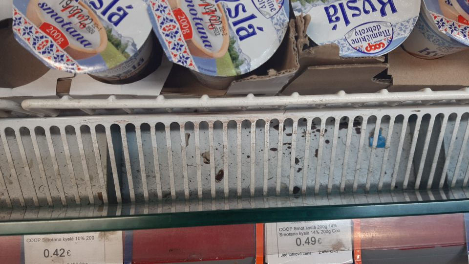 Značne znečistená chladiaca vitrínana mliečne výrobky v dolnej časti pod mriežkou prachom, zaschnutými zbytkami potravín (jogurt) a inými nečistotami.