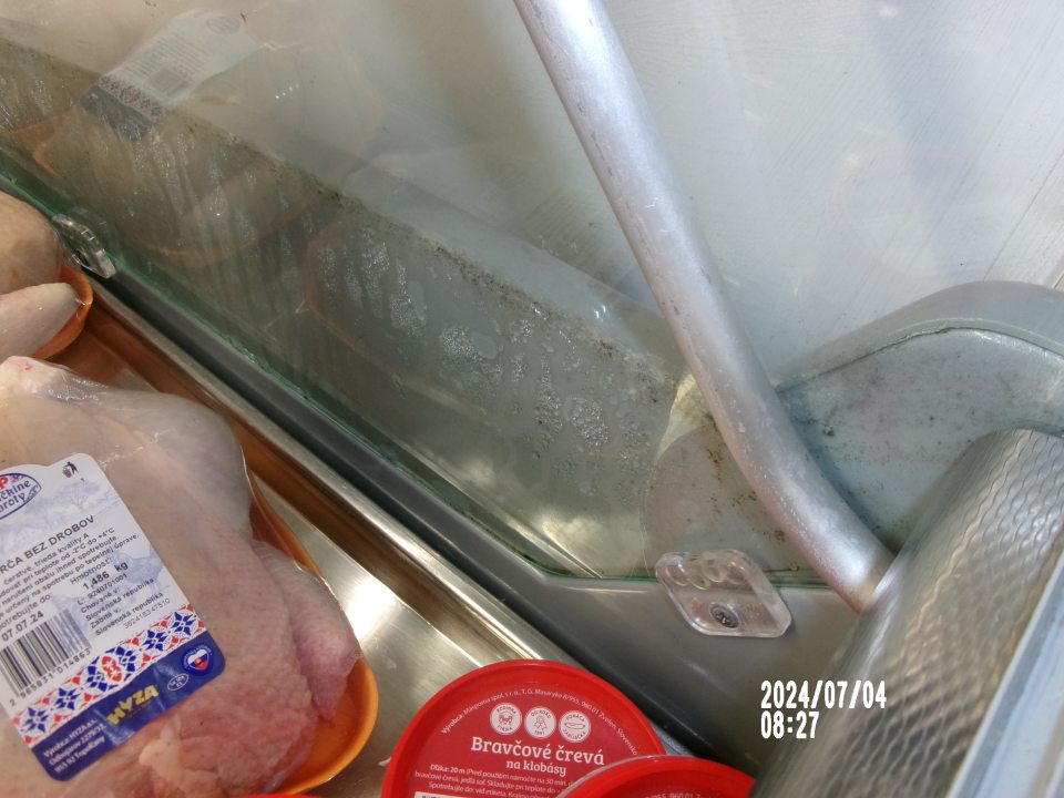 Zistená silne znečistená chladiarenská vitrína s uchovávanými mäsovými výrobkami, a to medzi bočnou sklenenou stenou a kovovou konštrukciou.
