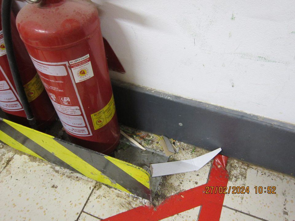 Znečistená podlaha prachom, nánosmi zaschnutej čiernej špiny, kúskami kartóna a rôznymi organickými nečistotami pri vstupných dverách do skladovacích priestorov. 