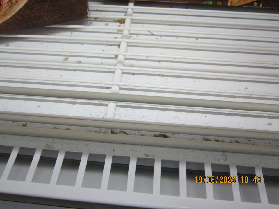 Prachom, organickými nečistotami a  zbytkami potravín  znečistená spodná časť pod mriežkami v  mraziacej vitríne na uchovávanie hlbokozmrazenej pizze na predajnej ploche.