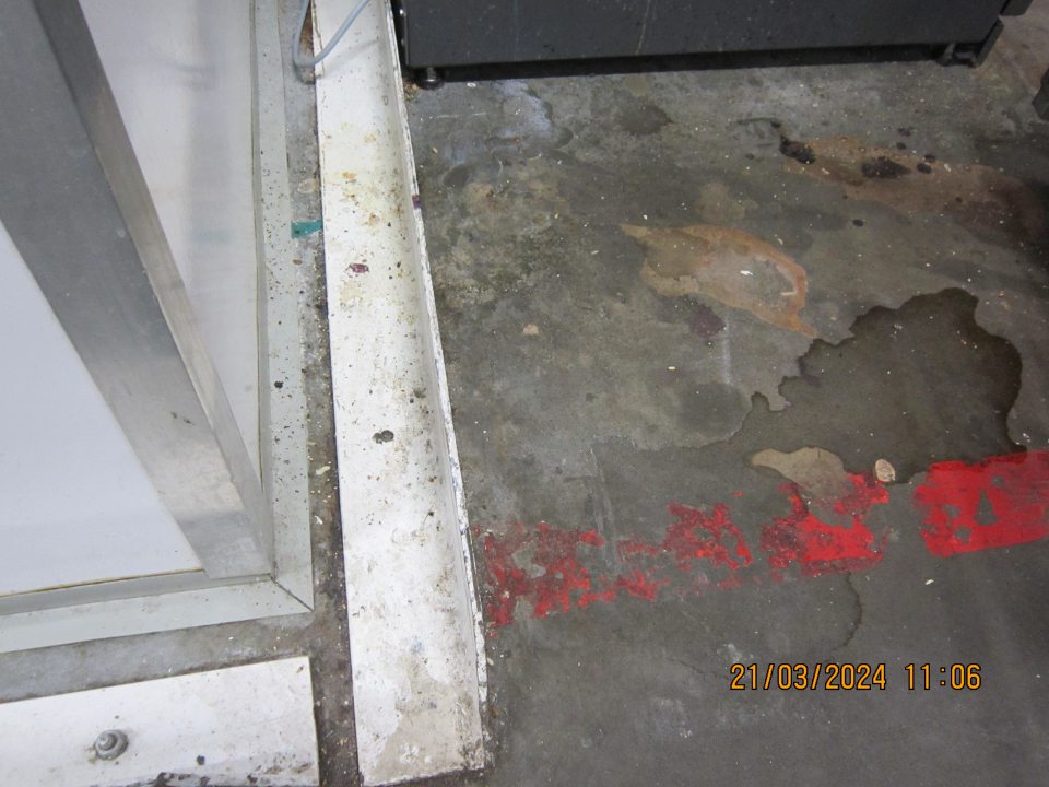 Organické nečistoty, zaschnutá špina, usadený prach na zemi za protinárazovou zábranou a podlaha znečistená zaschnutou tekutinou (zatečenou z potravín) v blízkosti chladiacej vitríny na uchovávanie odpisov v manipulačnej chodbe skladu smerujúcej k predajnej ploche.