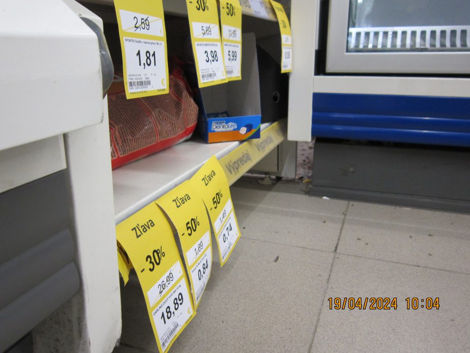 Znečistená podlaha v priestore medzi mraziacou vitrínou na uchovanie mrazených krémov a regálom so zľavneným tovarom zaschnutou čiernou špinou, prachom a kúskami papiera.