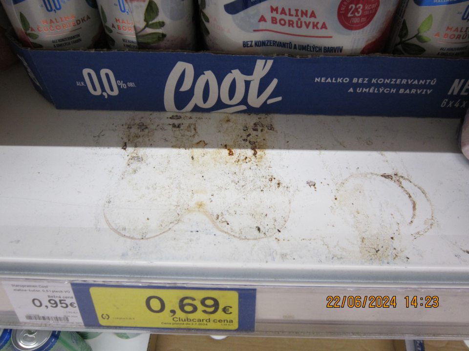 Znečistený regál na uchovávanie nealkoholických pív v plechovkách na predajnej ploche zaschnutými nečistotami z vytečeného obsahu.