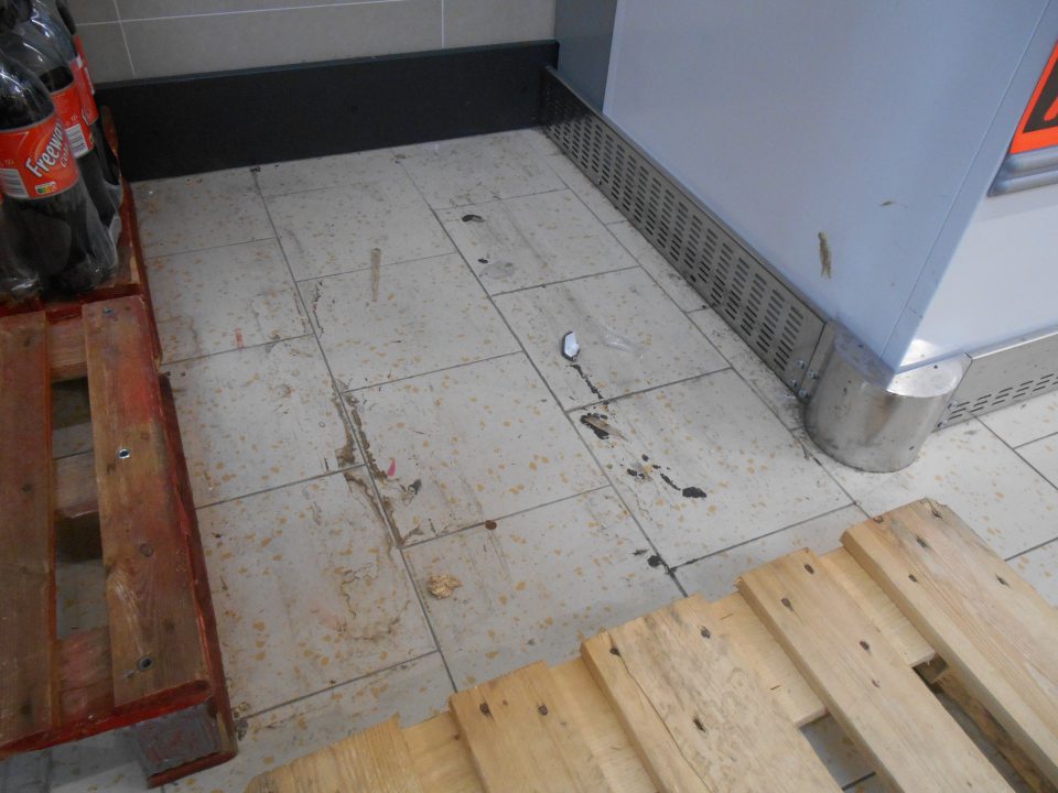 Znečistená podlaha na predajnej ploche prevádzky  pod paletami s nealko nápojmi pri mraziarenskej  vitríne s hlbokozmrazenými polotovarmi zaschnutou čiernou špinou, prachom, kúskami papiera a drobnými úlomkami z drevenej palety.