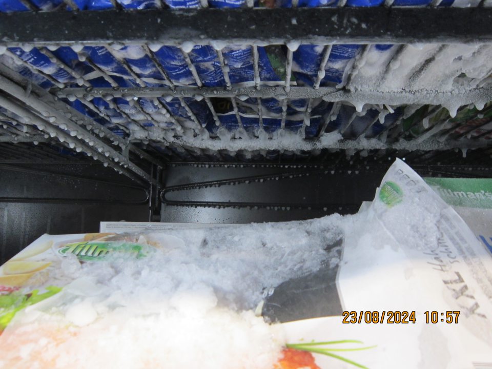 Zamrznuté kvapky vody na predeľovacích policových mriežkach a nadmerná námraza na kartónových baleniach znehodnoteného výrobku – hlbokozmrazený polotovar v poškodenom obale v mraziacej vitríne na predajnej ploche. 