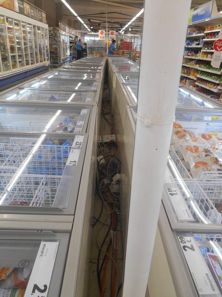 Podlaha znečistená prachom, kúskami papiera, kartónov a plastovými obalmi, neprehľadne zvinuté napájacie elektrické káble  v priestore medzi mraziacimi  boxami usporiadanými do dvojradu na predajnej ploche prevádzky.