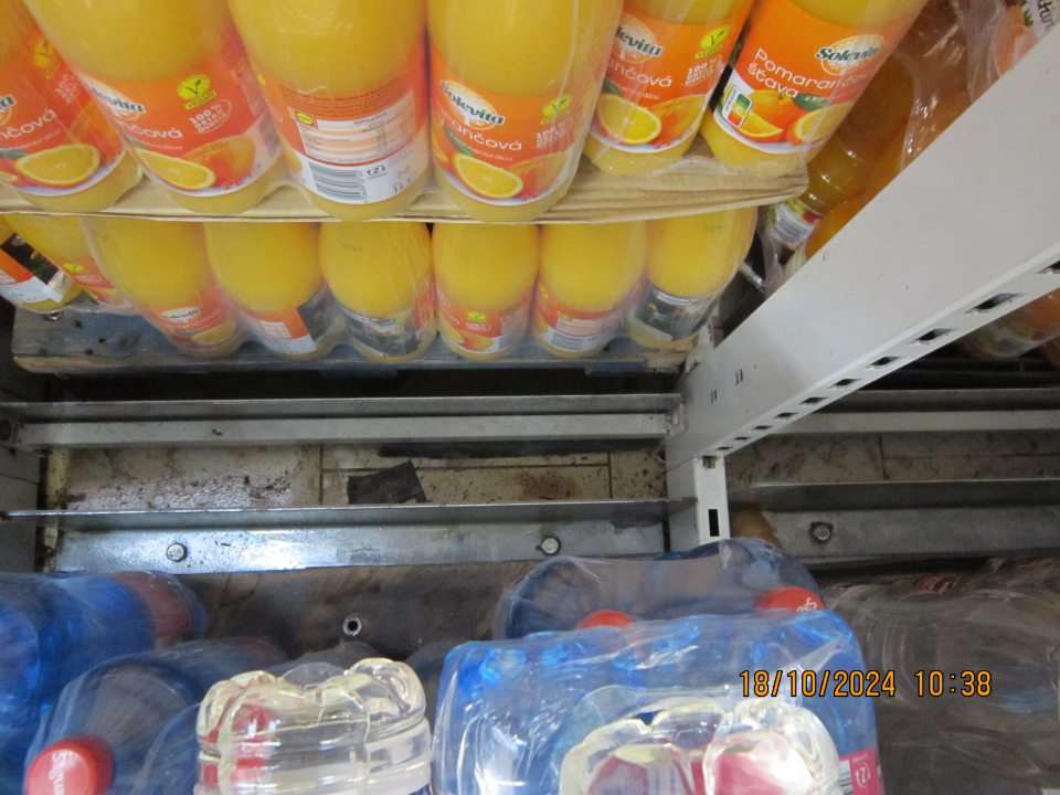 Podlaha v priestore medzi paletami s balenou minerálnou vodou a regálom so sladenými nápojmi znečistená prachom, kúskami plastov a vytečeným obsahom.