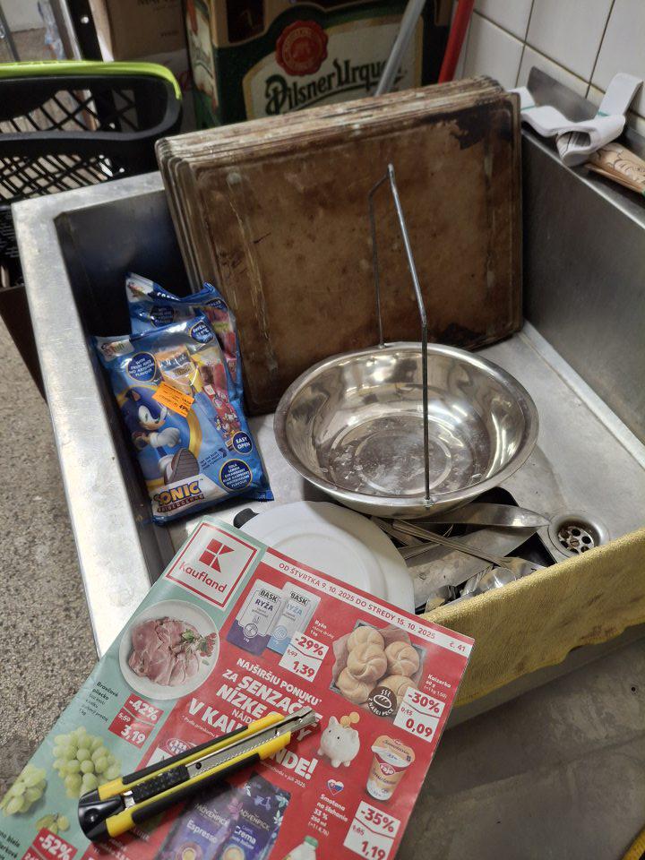 V nerezovom dvojdreze nahádzané plechy na pečenie, kuchynský riad zamestnancov, dvojdrez znečistený