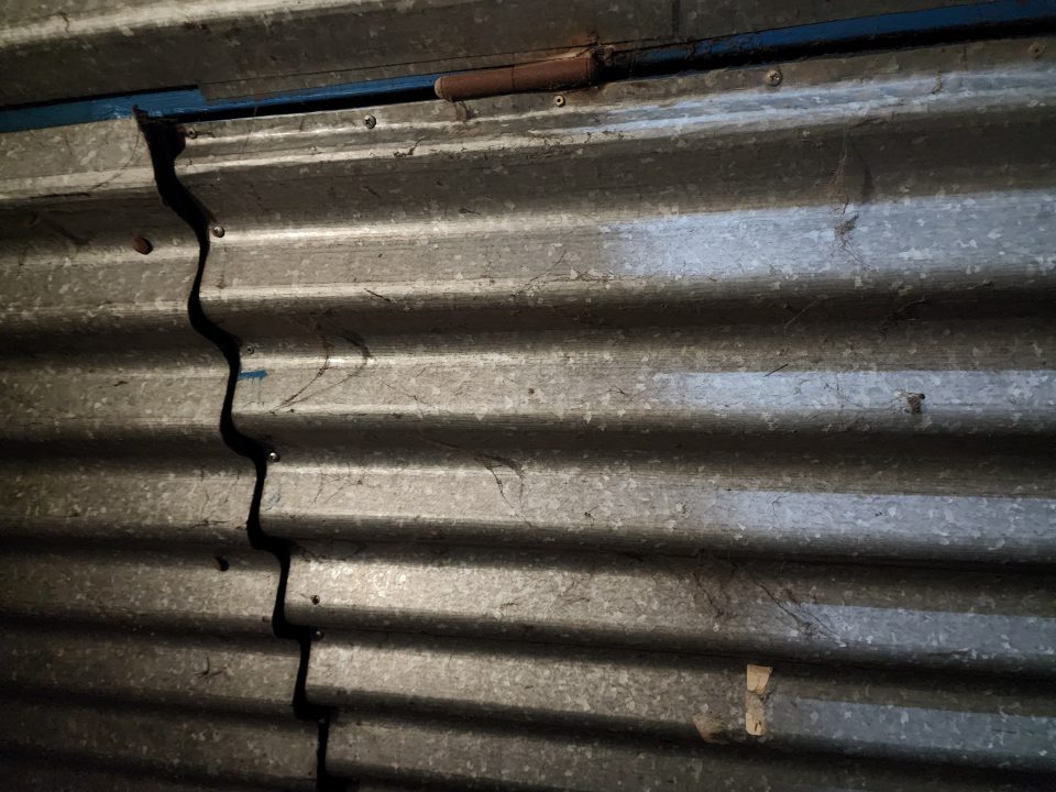  Vedľa príjmových dverí je stena prikrytá vlnitým plechom, na ktorom sú čierne hrubé pavučiny. 