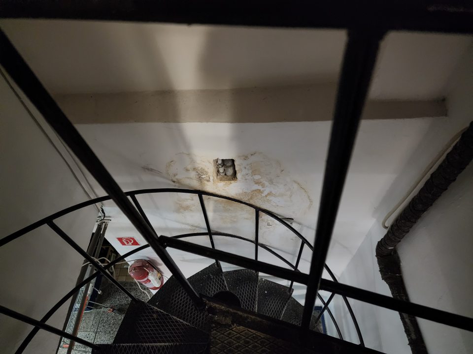 Silno zatečená stena s olupujúcou sa omietkou vedľa točitých kovových schodov vedúcich do suterénu.