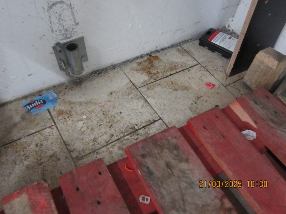 Podlaha znečistená nálepmi čiernej špiny a zbytkami obalového materiálu v priestoroch skladu v rohu pri príjmovej rampe v zázemí prevádzky.