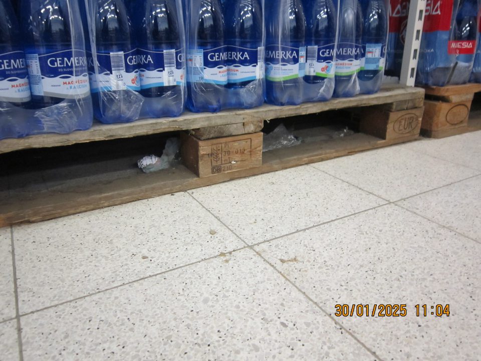 Nánosy prachu, nečistoty a zbytky obalového materiálu pod paletami s balenými minerálnymi vodami  na predajnej ploche prevádzky. 