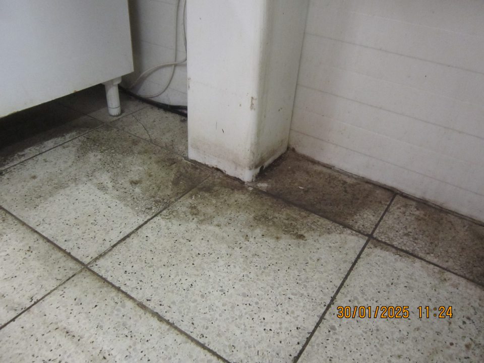 Podlaha pozdĺž steny v zázemí pultového predaja mäsa na predajnej ploche prevádzky znečistená nálepmi čiernej špiny.