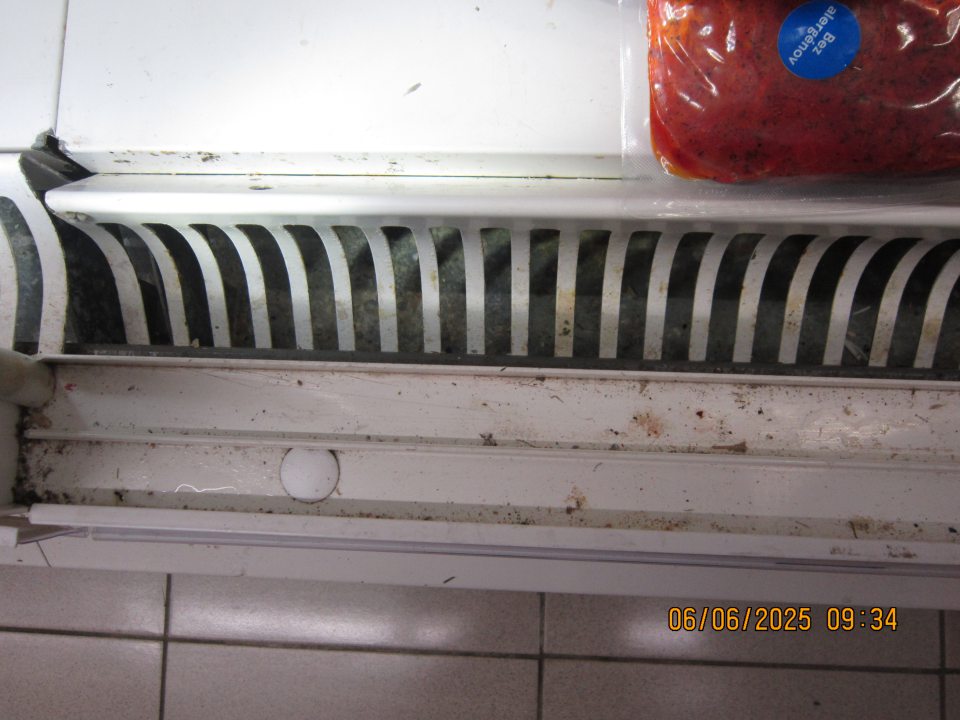 Silne znečistené mriežky, priestor pod mriežkami a  drážky zasúvacích dvierok v chladiarenskej vitríne na uchovávanie mäsových výrobkov– prach, drobné nečistoty a zaschnutá čierna nečistota.