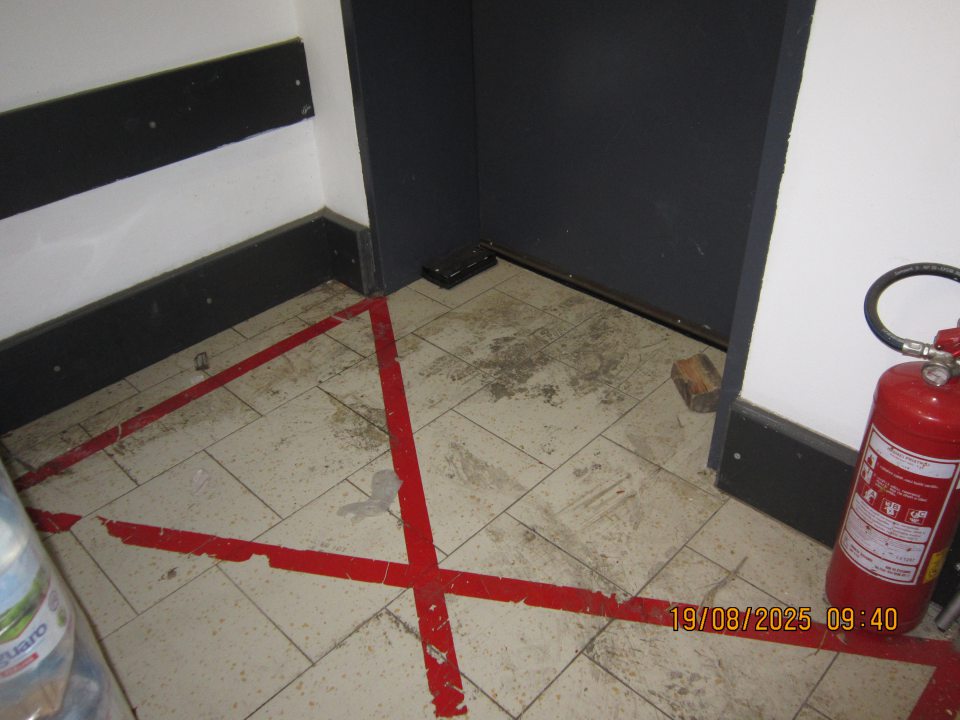 Vrstva čiernej špiny nalepená na podlahe v okolí dverí do vonkajšieho priestoru pri príjmovej rampe v  skladovacej časti prevádzky.
