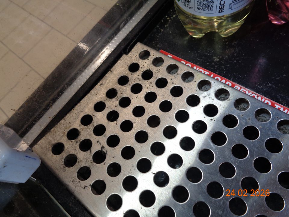 Znečistená kovová mriežka a priestor pod kovovou mriežkou na dne  chladiarenského zariadenia „Monster Energy“ na uchovávanie nealkoholických nápojov  na predajnej ploche hrubou vrstvou prachu a pavučinami.  