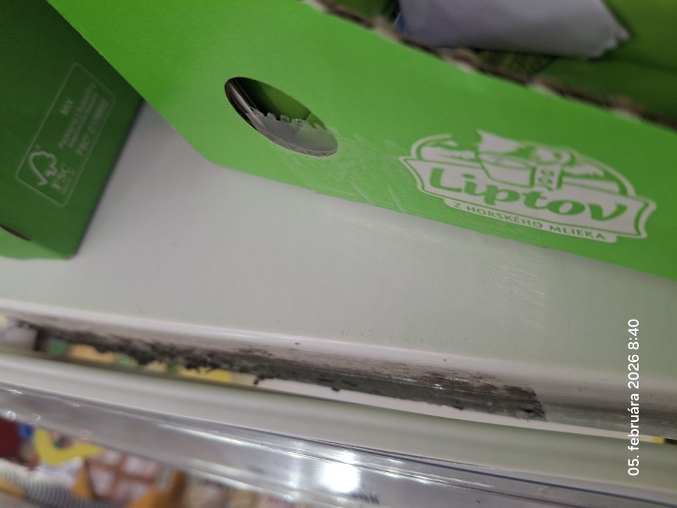 Na predajnej ploche znečistený regál v chladiacom zariadení na mliečne výrobky pod lištou na cenovky čiernymi nečistotami. 